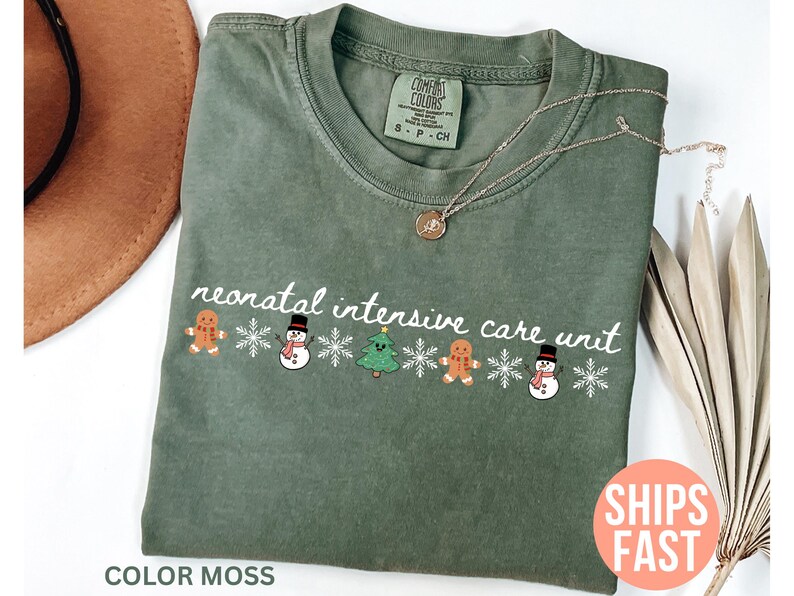 May include: Olive green t-shirt with the text "neonatal intensive care unit" and festive gingerbread men, snowmen, and Christmas trees. The shirt has a crew neck and short sleeves. The text "COLOR MOSS" is at the bottom.