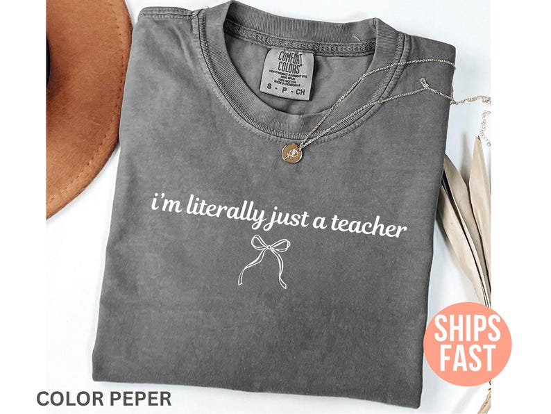 May include: A gray Comfort Colors t-shirt with the text "i'm literally just a teacher" and a bow graphic. A gold pendant necklace is draped on the shirt. The shirt is folded and has a vintage look.