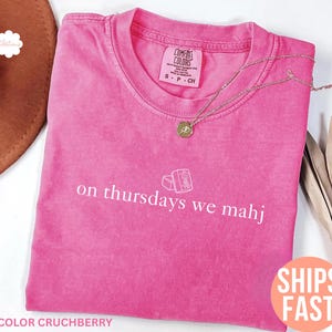 以下が含まれることがあります： ピンク色のComfort Colors Tシャツ。白い文字で「on Thursdays we mahj」と書かれています。 金色のネックレスがTシャツの上にあります。 Tシャツは無地で、クルーネックです。