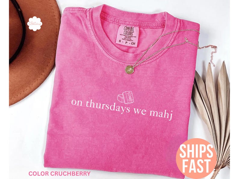 以下が含まれることがあります： ピンク色のComfort Colors Tシャツ。白い文字で「on Thursdays we mahj」と書かれています。 金色のネックレスがTシャツの上にあります。 Tシャツは無地で、クルーネックです。