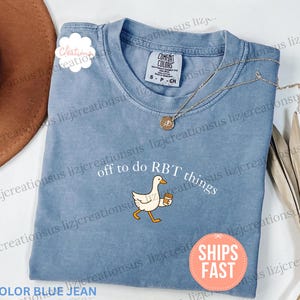 May include: A blue jean colored short-sleeve t-shirt featuring a graphic of a white goose holding a brown coffee cup. The text above the goose reads "off to do RBT things" in white lettering. The shirt has a crew neckline and a Comfort Colors label.