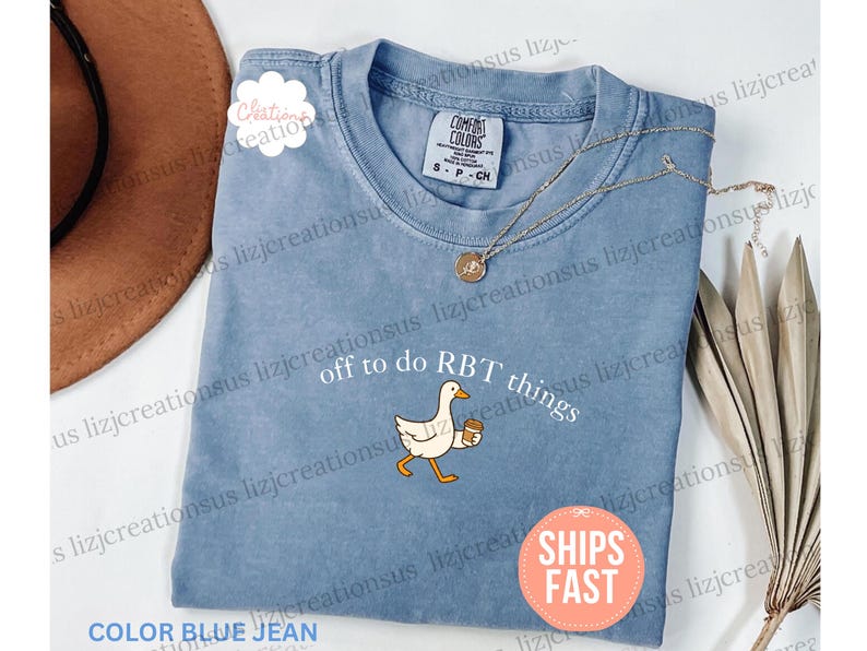May include: A blue jean colored short-sleeve t-shirt featuring a graphic of a white goose holding a brown coffee cup. The text above the goose reads "off to do RBT things" in white lettering. The shirt has a crew neckline and a Comfort Colors label.