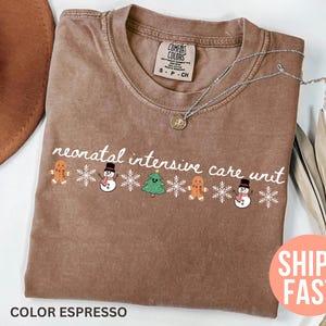 May include: Espresso-colored t-shirt with the words "neonatal intensive care unit" in white script. The shirt features a festive design of gingerbread men, snowmen, Christmas trees, and snowflakes. A silver necklace with a pendant is also visible.