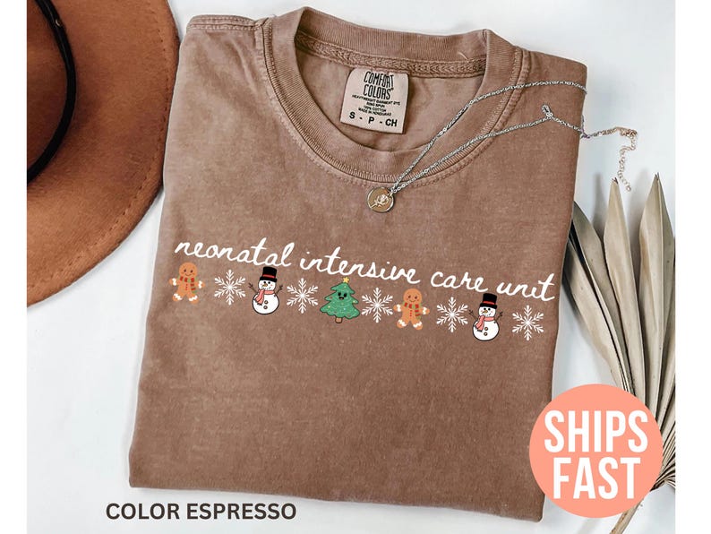 May include: Espresso-colored t-shirt with the words "neonatal intensive care unit" in white script. The shirt features a festive design of gingerbread men, snowmen, Christmas trees, and snowflakes. A silver necklace with a pendant is also visible.