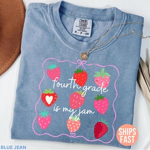 Könnte beinhalten: Ein blaues Jeans-T-Shirt mit Erdbeermuster und dem Text "fourth grade is my jam". Das Design zeigt einen rosa Rahmen mit mehreren Erdbeeren. Eine goldene Halskette liegt über dem Shirt.