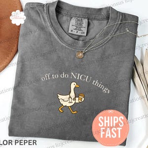 Puede incluir: Una camiseta gris carb&oacute;n de manga corta con un gr&aacute;fico de un ganso blanco que lleva un estetoscopio y sostiene una taza de caf&eacute;. El texto sobre el ganso dice off to do NICU things en letras blancas. La camiseta tiene una etiqueta Comfort Colors.
