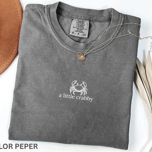 May include: A gray Comfort Colors t-shirt with a small white crab graphic and the text "a little crabby". A silver necklace with a round pendant rests on the shirt. A brown hat is partially visible.