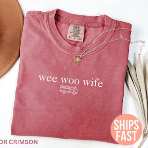 Può includere: T-shirt color cremisi con la scritta "wee woo wife" e l'illustrazione di un camper. Una collana d'oro con un ciondolo è appoggiata sulla maglietta. La maglietta è piegata su una superficie bianca.