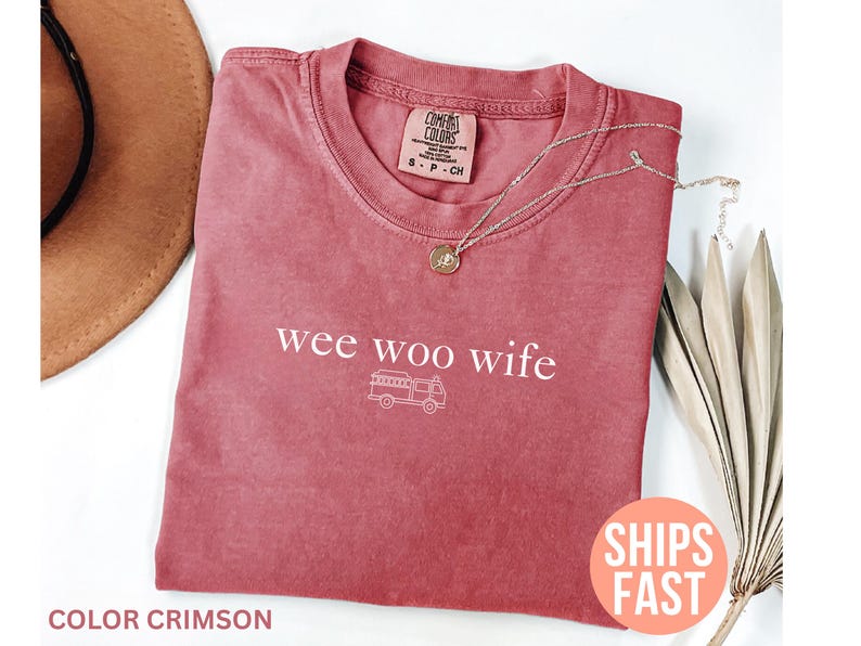 May include: Crimson-colored t-shirt with the words "wee woo wife" and a small illustration of a camper van. A gold necklace with a pendant rests on the shirt. The shirt is folded and placed on a white surface.
