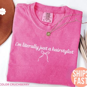 May include: A cerise pink t-shirt with the white text "I'm literally just a hairstylist". The shirt has a crew neck and a bow graphic. A gold necklace with a pendant is draped across the shirt.