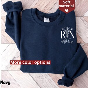 May include: Navy blue sweatshirt with a white graphic of a medical caduceus and the text "RN Ashley". The sweatshirt has a crew neck and long sleeves. More color options are available.