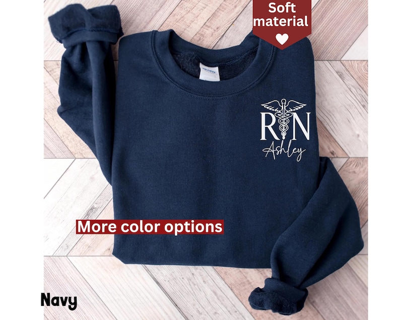 May include: Navy blue sweatshirt with a white graphic of a medical caduceus and the text "RN Ashley". The sweatshirt has a crew neck and long sleeves. More color options are available.