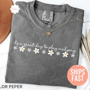 May include: A gray t-shirt with the phrase "it's a great day to play mahjong" in white script and daisy graphics. A gold-colored necklace rests on the shirt. The shirt has a small tag that says "S P CH".