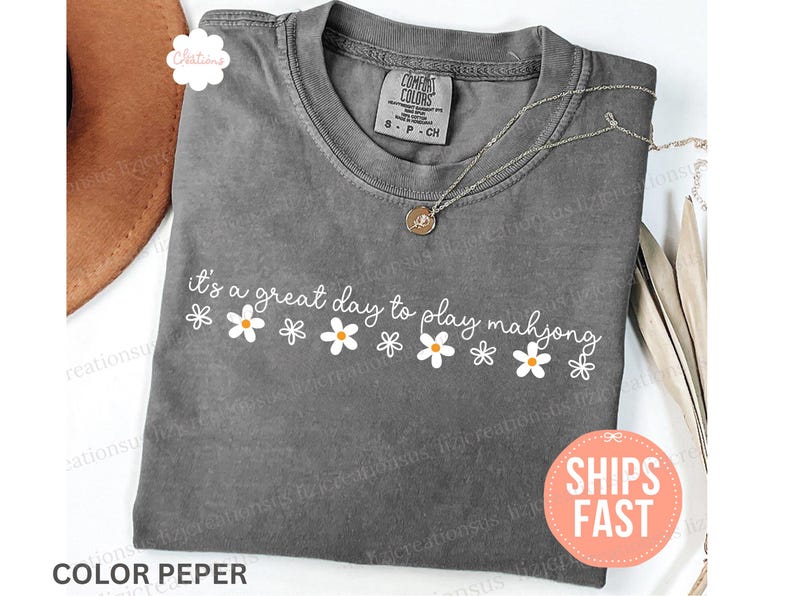 May include: A gray t-shirt with the phrase "it's a great day to play mahjong" in white script and daisy graphics. A gold-colored necklace rests on the shirt. The shirt has a small tag that says "S P CH".