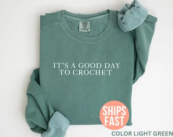 Puede incluir: Sudadera verde claro con el texto "IT'S A GOOD DAY TO CROCHET" en blanco. La sudadera está doblada, con las mangas y el cuello visibles. Una pegatina redonda con las palabras "SHIPS FAST" está en la parte inferior derecha.