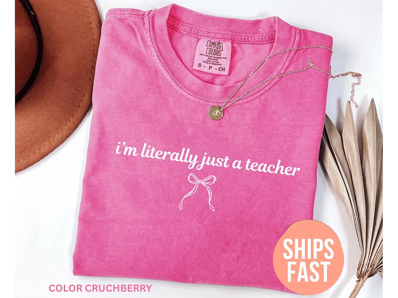 May include: A pink Comfort Colors t-shirt with the text "I'm literally just a teacher" and a bow graphic. A gold necklace with a pendant is draped across the shirt. The shirt is labeled "Color Crutchberry".