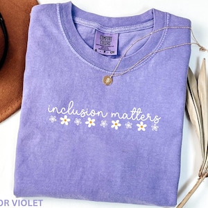 May include: A lavender Comfort Colors t-shirt with the phrase "inclusion matters" in white script and daisy graphics. A gold necklace and a brown hat are visible in the background. The shirt is a size small.
