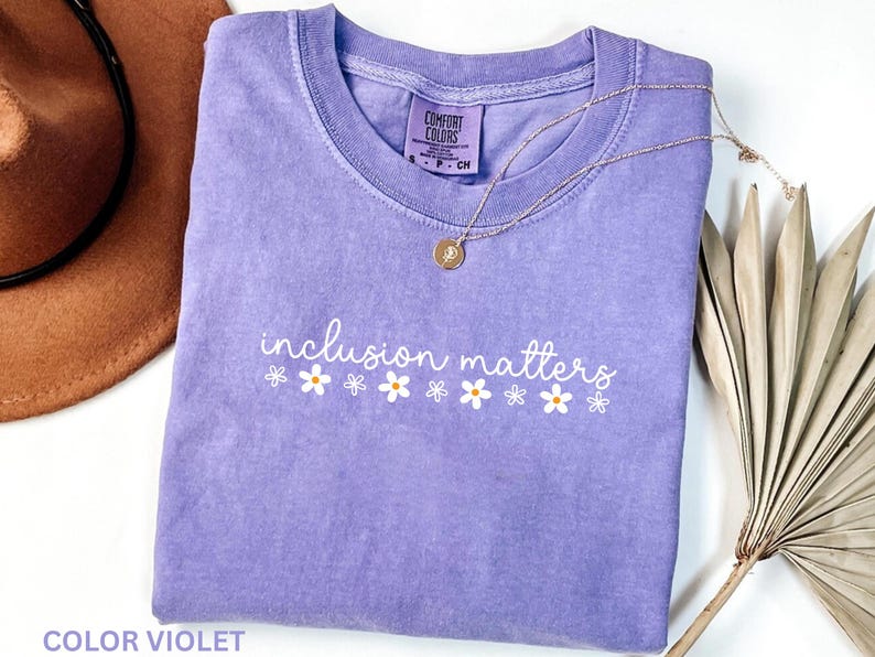May include: A lavender Comfort Colors t-shirt with the phrase "inclusion matters" in white script and daisy graphics. A gold necklace and a brown hat are visible in the background. The shirt is a size small.