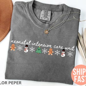 May include: A gray t-shirt with the words "neonatal intensive care unit" in white script. The shirt features a festive design with gingerbread men, snowmen, and a Christmas tree. A gold necklace rests on the shirt.