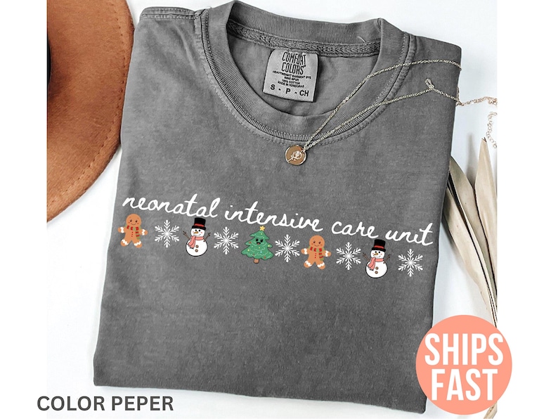 May include: A gray t-shirt with the words "neonatal intensive care unit" in white script. The shirt features a festive design with gingerbread men, snowmen, and a Christmas tree. A gold necklace rests on the shirt.
