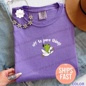 Può includere: Una t-shirt color violetta con una grafica raffigurante una rana verde che cavalca un'oca bianca dal becco arancione. Il testo sopra la grafica recita off to para things in lettere bianche. La maglietta presenta un girocollo e un'etichetta Comfort Colors.