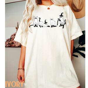 May include: Ivory-colored t-shirt featuring a black graphic of four cat-shaped ghosts. One ghost wears a witch hat. The shirt is a relaxed fit. The word "IVORY" is printed in orange at the bottom of the shirt.