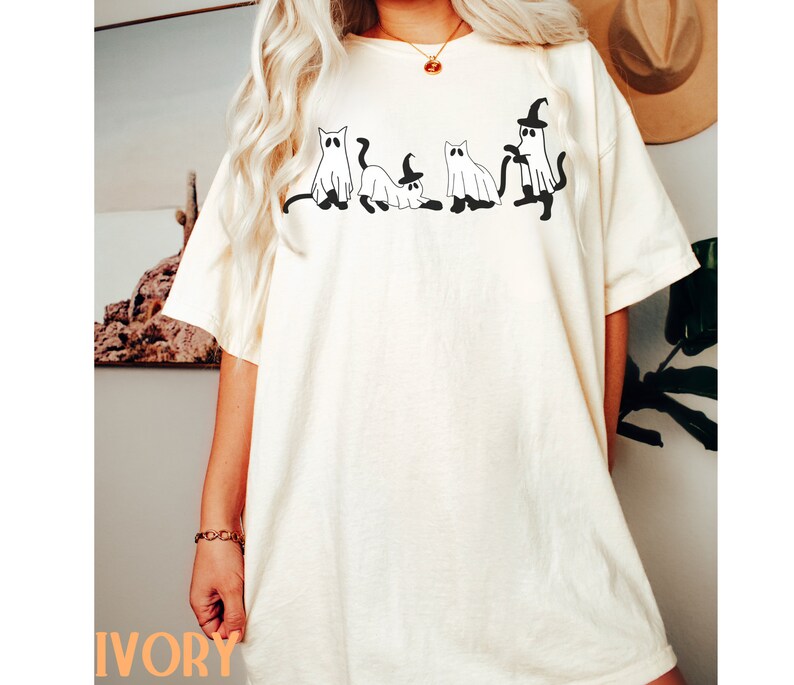 May include: Ivory-colored t-shirt featuring a black graphic of four cat-shaped ghosts. One ghost wears a witch hat. The shirt is a relaxed fit. The word "IVORY" is printed in orange at the bottom of the shirt.
