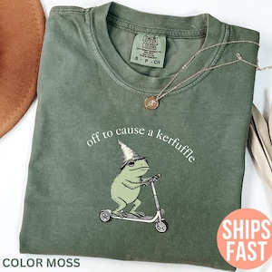 Könnte beinhalten: Moosgrünes T-Shirt mit einem Frosch auf einem Roller, der einen Zaubererhut trägt, und dem Text "off to cause a kerfuffle". Das T-Shirt hat ein Comfort Colors-Etikett. Eine goldene Halskette liegt über dem T-Shirt.