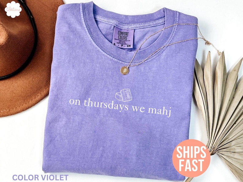以下が含まれることがあります： 「on thursdays we mahj」の白い文字がプリントされたラベンダー色のComfort Colors Tシャツ。 金色のペンダント付きネックレスがシャツの上にあります。 シャツは茶色の帽子と乾燥した葉の隣にあります。
