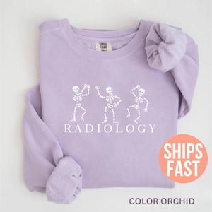 May include: A light purple sweatshirt with a crew neck. The front features a white graphic of three dancing skeletons above the word "RADIOLOGY". The bottom of the sweatshirt has the text "COLOR ORCHID".