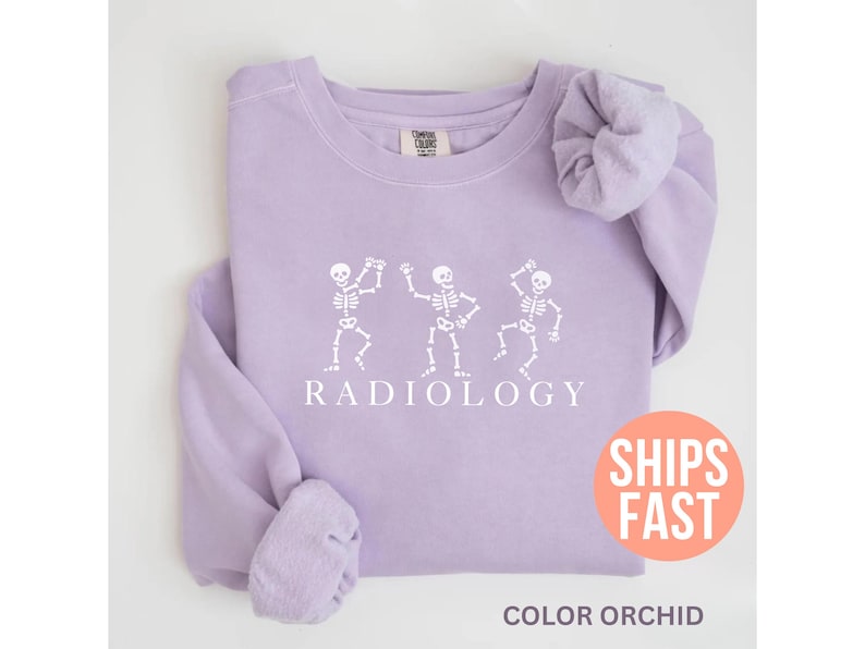 May include: A light purple sweatshirt with a crew neck. The front features a white graphic of three dancing skeletons above the word "RADIOLOGY". The bottom of the sweatshirt has the text "COLOR ORCHID".