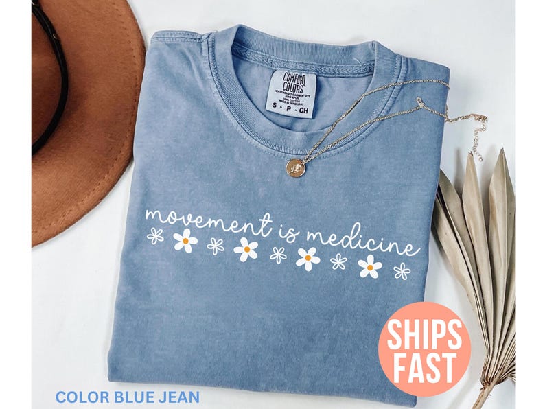 May include: Blue jean colored t-shirt with the phrase "movement is medicine" in white script and daisy graphics. A gold necklace with a pendant rests on the shirt. The shirt is folded, with a brown hat visible in the background.