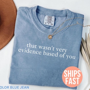 Könnte beinhalten: Blaues Jeans-T-Shirt mit dem weißen Schriftzug "that wasn't very evidence based of you". Eine goldene Halskette mit Anhänger liegt auf dem Shirt. Das Shirt ist gefaltet und hat ein Comfort Colors-Etikett.
