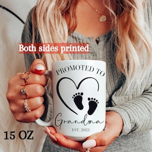 K&ouml;nnte beinhalten: Eine wei&szlig;e Keramiktasse mit schwarzem Text, der "Bef&ouml;rdert zur Oma Est. 2023" lautet. Die Tasse hat ein schwarzes Herz mit zwei Baby-Fu&szlig;abdr&uuml;cken darin. Die Tasse ist 15 Unzen.
