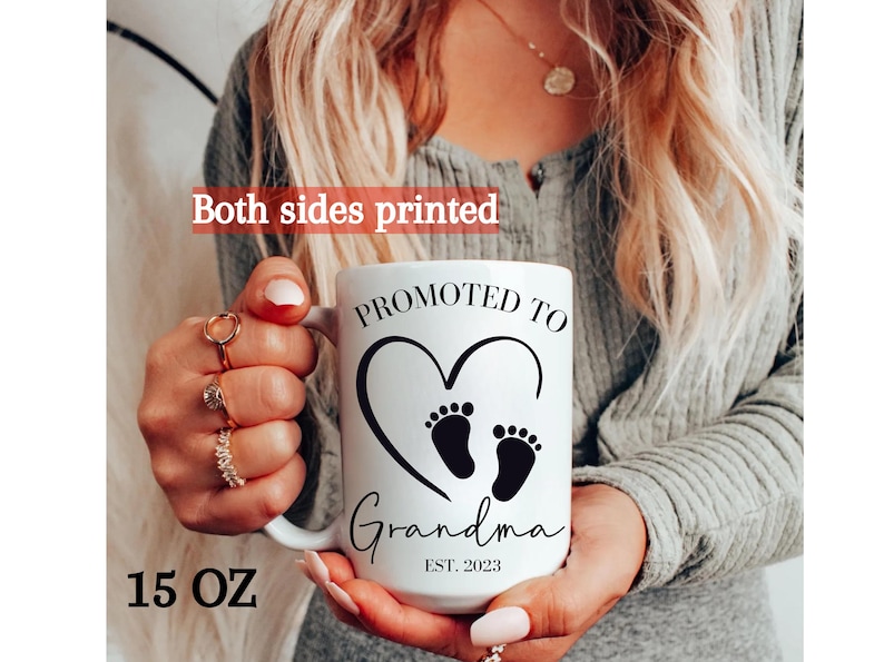 K&ouml;nnte beinhalten: Eine wei&szlig;e Keramiktasse mit schwarzem Text, der "Bef&ouml;rdert zur Oma Est. 2023" lautet. Die Tasse hat ein schwarzes Herz mit zwei Baby-Fu&szlig;abdr&uuml;cken darin. Die Tasse ist 15 Unzen.