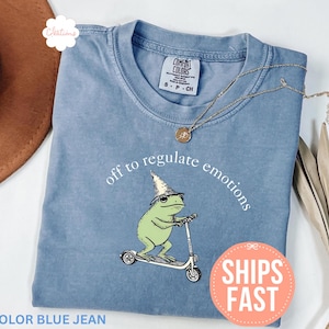 Puede incluir: Camiseta azul vaquero con un diseño de una rana verde con sombrero de mago en un patinete. El texto "off to regulate emotions" está arqueado arriba. Un collar dorado y una pegatina que dice "SHIPS FAST" también son visibles.