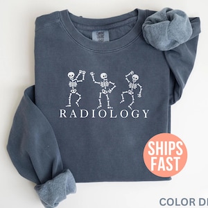 May include: A navy blue sweatshirt with the word "RADIOLOGY" printed in white, below three dancing skeleton figures. The sweatshirt has long sleeves and a crew neck. The text "SHIPS FAST" is on a red circle.