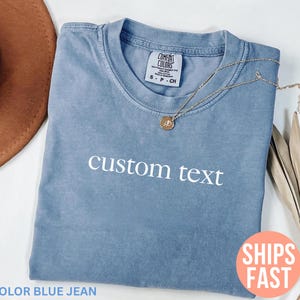 Op de afbeelding: Een blauw spijkerbroekkleurig t-shirt met de woorden "custom text" in het wit gedrukt. Een gouden ketting met een hanger rust op het shirt. Het shirt is opgevouwen en heeft een label met "Comfort Colors".