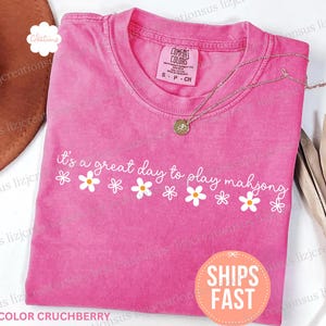 May include: A pink Comfort Colors t-shirt with the phrase "it's a great day to play mahjong" in white script, decorated with daisy graphics. A gold necklace rests on the shirt. The shirt is size S-P-CH.