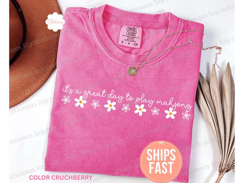 May include: A pink Comfort Colors t-shirt with the phrase "it's a great day to play mahjong" in white script, decorated with daisy graphics. A gold necklace rests on the shirt. The shirt is size S-P-CH.