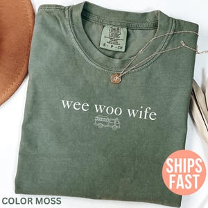 May include: A moss green t-shirt with the phrase "wee woo wife" and a fire truck graphic in white. A gold necklace with a pendant rests on the shirt. The shirt is folded on a white surface.