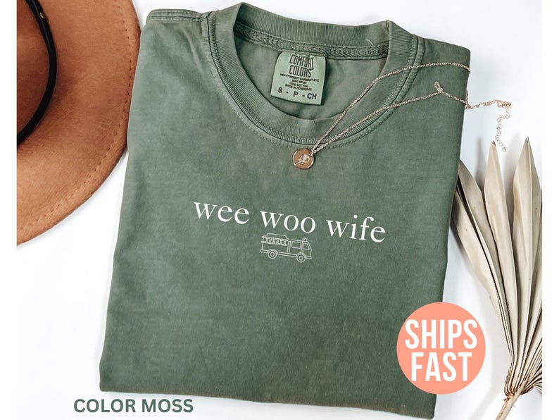 May include: A moss green t-shirt with the phrase "wee woo wife" and a fire truck graphic in white. A gold necklace with a pendant rests on the shirt. The shirt is folded on a white surface.