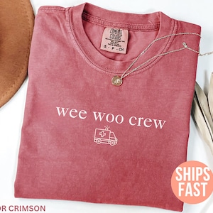 May include: Crimson-colored t-shirt with the text "wee woo crew" and an ambulance graphic. A silver necklace with a pendant rests on the shirt. The shirt has a Comfort Colors tag.