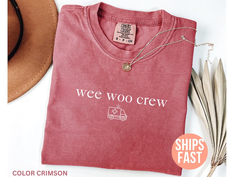 May include: Crimson-colored t-shirt with the text "wee woo crew" and an ambulance graphic. A silver necklace with a pendant rests on the shirt. The shirt has a Comfort Colors tag.