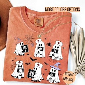 Puede incluir: Camiseta naranja quemado con un gráfico de fantasmas de dibujos animados sosteniendo radiografías, usando sombreros de bruja y gorras de enfermera. La camiseta tiene el texto "More Colors Options" y "Burnt Orange". Un collar dorado está drapeado sobre la camiseta.