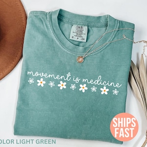 May include: Light green t-shirt with the phrase "movement is medicine" in white script and daisy graphics. A silver necklace with a pendant rests on the shirt. The shirt is folded and placed on a neutral surface.