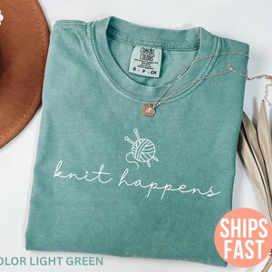 May include: A light green t-shirt with the words "knit happens" in white script, along with a graphic of yarn and knitting needles. A gold necklace with a pendant rests on the shirt. The shirt is folded and ready to wear.