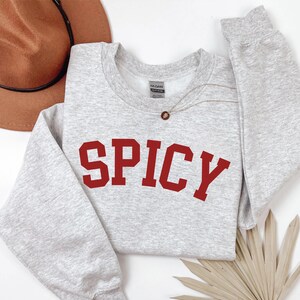 Peut inclure: Un sweat-shirt gris avec le mot "SPICY" imprim&eacute; en rouge sur le devant. Le sweat-shirt est fait d'un tissu doux et confortable et a une coupe d&eacute;contract&eacute;e.