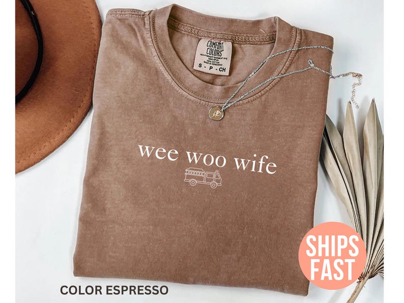May include: Espresso-colored t-shirt with the phrase "wee woo wife" and a graphic of a fire truck. The shirt has a crew neck and short sleeves. A silver necklace with a pendant is draped across the shirt. The shirt is folded on a white surface.