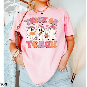 May include: Pink t-shirt with the words "Trick or Teach" in a retro font. The graphic features three cartoon ghosts, one reading a book, one with a pencil, and one with a jack-o'-lantern bucket. The shirt is a comfortable, casual style.